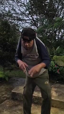 The video features a person outdoors in what appears to be a garden or yard. The person is wearing a grey and blue long-sleeved shirt, dark green pants, sunglasses, and a black cap, with a pink object appearing to be attached to the back of the cap. The person is standing on stone steps and briefly approaches the camera, then turns away and walks down the steps. The environment includes trees, bushes, and some lighting.