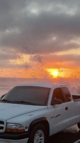 The video shows a scenic view of a sunset over the ocean, with waves crashing against rocks. A white pickup truck is parked on the side of the road, and a white SUV is driving past. The sky is filled with dramatic clouds in shades of orange, pink, and gray. The sun is a bright orb, casting a golden glow on the water. In the distance, a long pier extends out into the ocean. The road appears to be wet, possibly from recent rain or sea spray. The overall mood is peaceful and serene, with a touch of drama from the crashing waves and vibrant sunset.
