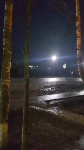 The video captures a nighttime outdoor scene in Krasnogorskoye, Russia, featuring a street and surrounding area. It opens with a close-up view of two tree trunks on the left and another slender tree in the center foreground. The ground in the foreground is dark and appears wet, possibly from rain or melted snow. The camera then slowly pans to the right, revealing a broader view of the street. A bright street lamp stands prominently in the mid-ground, casting a strong light that creates reflections on the wet asphalt and puddles on the ground. To the right of the street lamp, a yellow triangular pedestrian crossing sign, depicting a walking figure, is clearly visible on a pole. A person can be vaguely discerned standing near this sign. In the distance to the left, a building with illuminated windows is visible. A small, dark vehicle briefly passes beneath the street lamp. The scene is dark, damp, and dimly lit by artificial light. Throughout the video, faint, indistinct speech is audible.