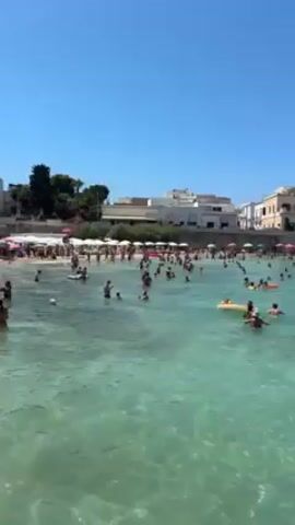 A crowd of people stand and wade in shallow, clear water. White beach umbrellas and lounge chairs are visible on the sandy beach in the background. Most people are wearing swimsuits. One man is walking and appears to be bending forward slightly. Some people are conversing.