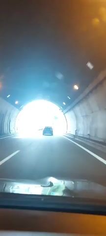 The video begins with a view from inside a vehicle traveling through a tunnel. The tunnel exit is visible ahead, bright with daylight. As the vehicle exits the tunnel, it merges onto a highway. The highway is flanked by trees and rolling hills on either side. The sky is overcast. Traffic can be seen on the highway, including other cars and trucks. The video shows the vehicle traveling on the highway for a short period before ending.