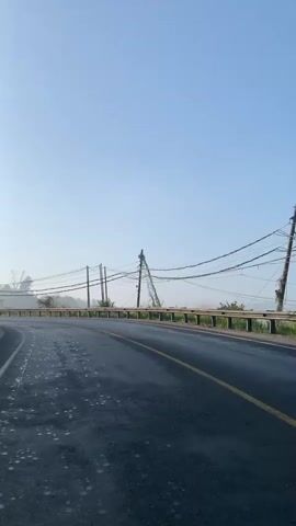 The video shows a road curving to the left, with a guardrail on the right side. There are telephone poles and wires running alongside the road. The sky is clear and blue, and there is some mist or fog in the distance. The road appears to be wet, possibly from rain or dew. The overall scene suggests an outdoor setting, possibly in a rural or mountainous area, during the morning. There are no people or animals visible in the video.