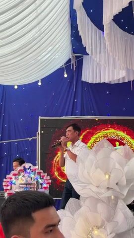 A man in a white, patterned short-sleeved shirt and dark pants is singing into a microphone, holding it close to his mouth and occasionally gesturing with his hands. He is positioned in front of a large screen displaying a dynamic red and yellow fiery pattern. Behind him, a blue fabric backdrop adorned with small lights creates a festive atmosphere. To his right, large white decorative flowers, resembling lotus blossoms, are prominent. Another man is partially visible behind a white decorative stand with small, clear containers, and a third man's face is seen in the foreground, looking to the left. The scene suggests a performance at a celebration or event in Tay Son Commune, Vietnam.