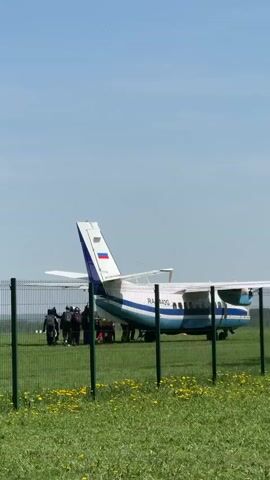 The video shows a small, blue and white aircraft, registered as "RA-4421G," parked on a grassy field. A group of people, mostly clad in dark clothing, stand near the plane, seemingly preparing for something. The area is fenced off with a green chain-link fence. Yellow dandelions dot the grassy foreground. The sky is clear and blue. The plane's propeller is blurred.