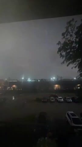This is a video of a stormy sky over a parking lot with cars. The sky is dark and foggy, with lightning illuminating the distance. The parking lot is wet, suggesting that it has been raining. There are several cars parked in the lot, including a white SUV and a few sedans. In the background, there are buildings with lights on. A tree is visible on the right side of the frame. The overall atmosphere is dark and moody, with the lightning adding an element of drama.