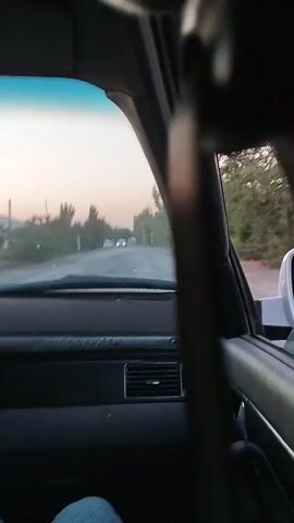The video shows a first-person perspective from inside a car driving down a dirt road. The sun is setting, casting a warm glow over the scene. Trees line both sides of the road, and there are houses visible in the distance. Two white cars are ahead of the car the video is being filmed from. The camera is focused on the road ahead, and the dashboard of the car is visible in the foreground. The video is shaky, indicating that the car is moving.