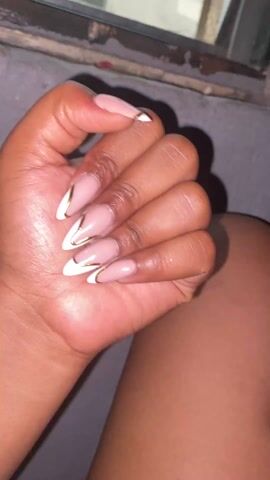 A person with tanned skin holds their left hand, showing manicured nails in an almond shape, with a white tip outlined in gold. The nails are clean and appear glossy. The person is sitting, with their thigh visible, and holding a smartphone in their right hand. The background shows a window with a metallic frame and a grey wall. The overall lighting appears dim.