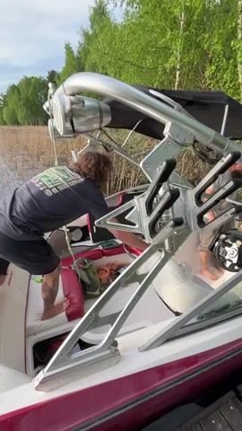 Two people are in a motorboat docked near a wooded area. The boat has a white interior with red accents and a large metal tower structure with speakers. One person, wearing a black shirt and shorts, is on the left side of the boat. The other person, wearing a light-colored shirt and dark pants, is near the driver's seat. They seem to be discussing something. The camera angle is slightly low, showing the boat from the side and focusing on the people inside. There is a wooden dock visible on the right side of the frame.