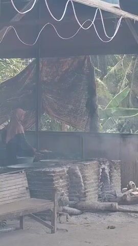 A woman wearing a hijab is preparing food over an open fire in an outdoor setting. She is standing and appears to be stirring or tending to food in a large pot or pan. The fire is contained within a brick structure that looks like a makeshift oven or cooking station. Smoke is rising from the fire, obscuring some of the background. In front of the cooking area, there is a wooden bench. The environment appears to be rustic, with trees and foliage visible in the background. The overall impression is of a traditional or rural cooking setup.
