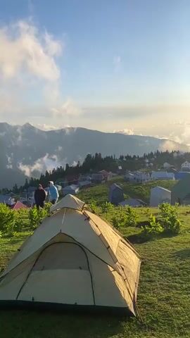This is a vertical video clip showcasing a picturesque mountain landscape in Gomismta, Georgia. The scene is bathed in the warm glow of the setting sun, casting long shadows across the grassy slopes. Fluffy clouds drift across a clear blue sky, partially obscuring the sun and creating a beautiful, ethereal atmosphere.

In the foreground, a tan tent is pitched on a verdant hillside, suggesting camping or an outdoor adventure. Further down the slope, a small village of houses with red and green roofs is nestled amidst the rolling hills. Some of these structures appear to be traditional mountain homes.

Several people are visible throughout the clip. In the mid-ground, two individuals stand close together, seemingly enjoying the view. Further to the right, a group of people are gathered, with some appearing to be having a picnic or engaging in conversation. Another person is seen walking away from the camera in the distance.

The overall mood is serene and tranquil, capturing the beauty of a mountain sunset and the simple pleasures of being in nature. The time of day is clearly evening, given the low angle of the sun and the warm lighting. The weather appears to be pleasant, with a mix of sun and clouds. The location cues, such as the style of houses and the mountainous terrain, strongly suggest the rural highlands of Georgia. There is no visible text in the video.