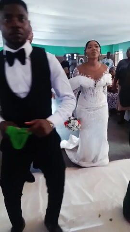 The video shows a bride and a man, presumably the groom, walking through what appears to be a wedding reception venue. The bride is wearing a long white lace gown with a bouquet in her hands. The man is dressed in a black vest and bowtie.  Other people are also visible in the background, some seated at tables, possibly guests at the wedding. White confetti is being thrown as the couple walks through the room. The scene is brightly lit, and the walls of the room appear to be painted a light green.