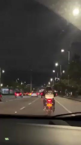 The video shows a night time street scene from the inside of a car. Other vehicles are driving on the same street, including a motorcycle with a delivery box on the back, and a car with its lights flashing. Streetlights are present on both sides of the road. The weather appears to be clear.