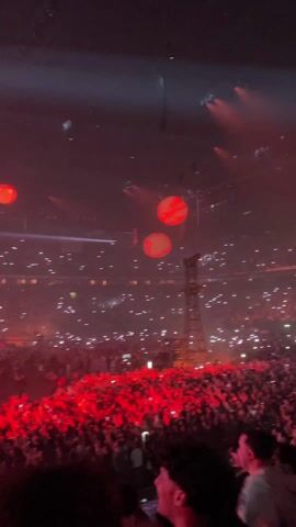 The video captures a live concert in a large arena packed with concertgoers. A stage is visible in the distance, illuminated by spotlights and pyrotechnics. Red balloons float over the audience, adding to the spectacle. The crowd is dense, with many people holding up their phones to record the performance. The lighting is intense and dynamic, creating a vibrant atmosphere.
