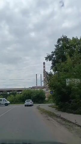 A white sedan turns a corner on a paved road. There is a pedestrian crossing sign on the right side of the road. In the background, there are industrial buildings and smokestacks, along with trees and power lines. The sky is cloudy.