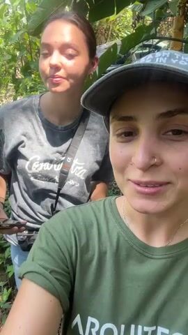 The video features two women in a lush, tropical environment. The woman in the foreground wears a green t-shirt with "ARQUITECT" printed on it and a grey baseball cap. She has a nose piercing and smiles at the camera, waving at one point. The second woman, slightly behind, wears a grey Coca-Cola t-shirt, jeans, and a camera strap. She briefly holds up a phone. The background shows dense greenery, including banana trees with hanging bananas. There are also glimpses of a concrete structure amidst the foliage, possibly the remnants of a building or foundation. The overall impression is of an outdoor exploration in a tropical setting.