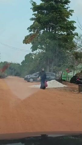 A person wearing a black hijab and black abaya walks across a dirt road, carrying a red bag. A grey car is parked on the left side of the road. In the background, there are trees, a green building, and a white car. The weather appears to be overcast.