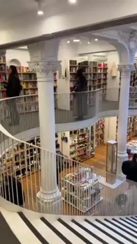 The video shows the interior of a multi-level bookstore, viewed from an elevated position on a staircase with black and white striped steps. The bookstore is busy with customers browsing the bookshelves on multiple levels. White pillars and ornate arches support the upper levels, creating an open and airy atmosphere. The store has a modern but classic look with dark wood floors, white shelving, and visible books on display. People are moving around on all levels, examining books and products on display tables. Natural light comes in through large windows. The overall impression is of a vibrant and well-maintained bookstore environment.