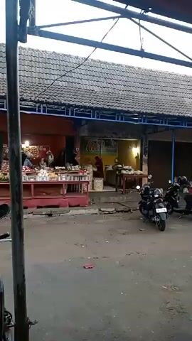 The video shows an outdoor market scene. There are several stalls with awnings, and people are visible behind the stalls and walking through the area. Various goods are displayed on tables and in baskets. Several motorcycles are parked in front of the stalls. The ground is paved with asphalt. The overall atmosphere appears to be a busy marketplace.