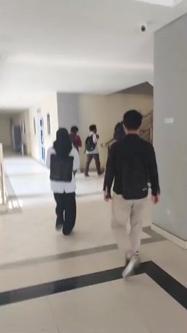 The video shows a hallway with several people walking. In the foreground, a person wearing a black jacket and light-colored pants walks away from the camera. To the left, another person in a white shirt and black pants, also wearing a backpack, walks away from the camera. Further down the hallway, several other individuals are visible, some walking and some near a staircase. The hallway appears to be inside a building, with tiled floors and a light-colored wall. There is a sign hanging on the wall to the left. The lighting is bright, suggesting it is daytime.