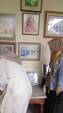 The video depicts two women exploring the interior of a historic building. Initially, one woman, dressed in a white top, light-colored hijab, and light-colored jeans, examines framed photographs on a wall. Subsequently, another woman, wearing a white top, black hijab, and black pants, is shown walking past a table with framed photos. The camera then follows this woman as she walks along a long corridor lined with antique furniture.