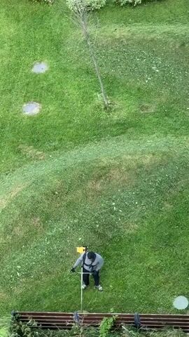 The video captures an aerial view of a person mowing a grassy slope with a string trimmer. The person is wearing a gray jacket, dark pants, and a yellow backpack. They are positioned on the slope, holding the trimmer and actively engaged in cutting the grass. The surrounding environment is characterized by a lush green lawn, with a thin tree and two circular lighter patches visible further up the slope. A wooden bench is situated at the bottom of the frame, with overgrown foliage in front of it. The footage appears to be shot from a high vantage point, looking down on the scene.
