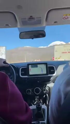 The video shows the interior of a car, specifically the front passenger area, from the perspective of someone in the back seat. The car is moving down a road on a sunny day. In the background, there's a mountain with a snow-covered peak. The driver, wearing a maroon shirt, is visible in the driver's seat, holding the steering wheel. To their right, the front passenger, wearing a white hoodie, is looking down at a mobile phone. The dashboard features a digital navigation screen displaying a route. Speed limit signs are visible on the side of the road.