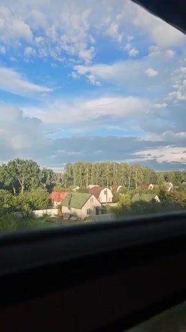 The video shows a rural landscape viewed through a window. The sky is blue with scattered white clouds, and a faint rainbow is visible. Below the sky, there are several houses with green and red roofs, surrounded by trees and greenery. The perspective suggests the viewer is in a moving vehicle.