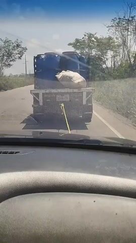 The video is taken from inside a vehicle driving behind a small, dark-colored flatbed truck. The truck is carrying multiple large, blue plastic barrels and a large white sack. The road appears to be a paved rural road, with vegetation on either side. Telephone poles are visible along the road. The sky is bright and sunny with scattered clouds. The windshield of the car is dirty in places. The dashboard is grey. The vehicle is in motion, following the truck. The license plate of the truck is partially visible, starting with "A9A0". A yellow tow strap is attached to the back of the truck. The camera is relatively stable, with minor movements due to the vehicle's motion.