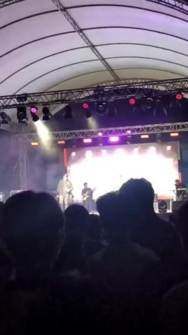 The video shows a live concert with a band performing on a stage. The stage is lit with colorful lights that change from red to purple to green and back to red. A crowd of people is visible in the foreground, silhouetted against the stage lights. Some members of the band can be seen on stage, playing instruments and interacting with the audience. The stage has a large backdrop with a logo that appears to say "northwest". The concert is taking place outdoors under a large tent-like structure.