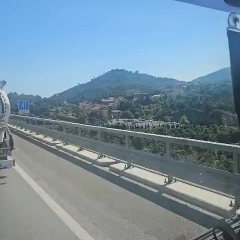The video shows a view from a vehicle driving on a bridge on a sunny day. In the distance, a town is nestled amidst a hillside, crowned by a large, fortified building on the summit. The town features various buildings, including houses, a church with a bell tower, and a parking lot filled with cars. The hillside is covered in lush greenery. The bridge has metal guardrails and a white lane marker. The sky is clear and blue.