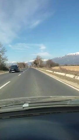The video shows a car driving on a two-lane highway on a clear day with a bright blue sky and some wispy clouds. The road is surrounded by dry, golden fields and sparse trees, some of which appear bare. In the distance, a mountain range capped with snow is visible. A yellow car passes in the opposite direction. The camera is positioned inside the car, looking out through the windshield. There is music playing.