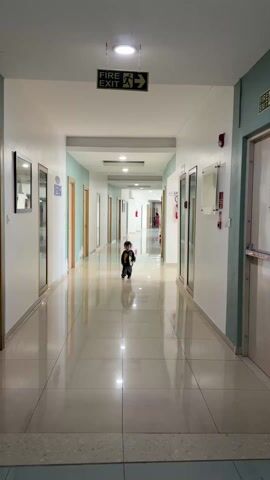 A toddler runs down a long, brightly lit hallway with shiny, reflective tile floors. The walls are painted white and pale blue and lined with doors and glass display cases. A "FIRE EXIT" sign hangs from the ceiling. In the distance, a woman stands near the end of the hall. The child is wearing dark clothing and runs towards the camera, giggling.
