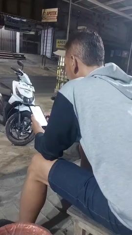 A man sits on a low wooden stool outside at night, facing a white and black scooter. He is wearing a grey hooded sweatshirt with black sleeves and dark blue shorts. He holds a smartphone in his left hand and appears to be looking at it. His right arm is bent, and his hand is resting on his leg. The scooter is parked to his left. In the background, there are buildings with signs that read "APOTEK". A road is visible behind the scooter, and a vehicle with its headlights on is driving by. The ground appears to be paved. The lighting suggests it is evening or nighttime.