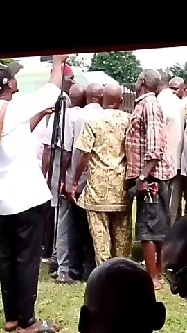 The video shows a group of men standing in a line outdoors. A man in a white shirt and black pants with a red cap is at the front of the line, holding what appears to be a rifle. He is gesturing with his left hand. The other men are mostly bald and are facing away from the camera, looking towards the man with the rifle. They are dressed in casual clothing, including a man in a patterned tunic and pants, and another in a plaid shirt and shorts. The environment appears to be grassy with some trees and a white structure in the background. The overall scene suggests a gathering or assembly of some kind.