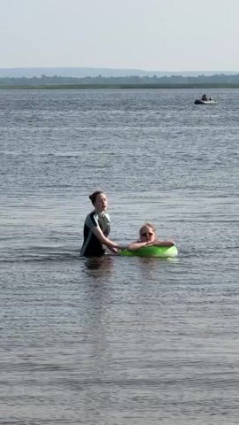 This media file shows two individuals in a body of water, likely a lake, on a bright, clear day. The setting is a vast expanse of water with a distant shoreline featuring trees and hills, suggesting a natural outdoor environment. The water appears calm and the sky is a pale blue, indicating fair weather.

In the foreground, a person wearing a black and teal swimsuit stands in the water, holding onto a green inflatable tube. This person is looking towards the camera with a slight smile. Inside the tube, another person, who appears younger, is seated and also looking towards the camera, wearing sunglasses. Both individuals seem relaxed and are enjoying a leisurely time in the water.

In the background, a small motorboat is visible on the water, indicating recreational activity is taking place in the broader area. The overall mood conveyed is one of summer enjoyment and outdoor recreation. No visible text is present in the media file. The time of day appears to be late morning or early afternoon due to the bright lighting. The context provided suggests the location is Krasnyi Bor, Russia.