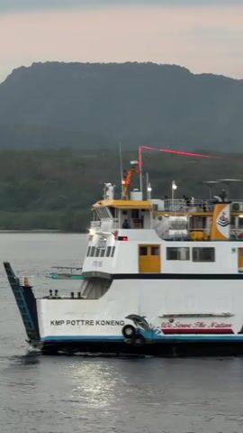 A ferry named KMP POTTRE KONENG is shown from its starboard side sailing on calm water. The ferry is white with yellow accents and has multiple decks. People are visible on the upper decks, some standing and some appearing to be seated. A red laser light beam extends across the top of the ferry. The ferry has the DLU Ferry logo and the text "We Serve The Nation" visible on its hull. In the background, a green, tree-covered hill or mountain rises from the water. The sky is overcast, suggesting dawn or dusk.