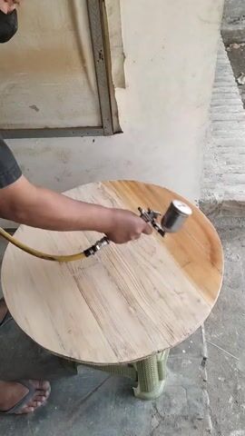A person's hands are visible as they spray a round wooden tabletop with a spray gun. The tabletop is light-colored and has visible wood grain. The spray gun is connected to a yellow hose, and the person is holding it with their right hand. The tabletop is resting on a green plastic stand. The background appears to be an outdoor or workshop setting with a textured wall and some bricks. The person is wearing a black shirt and dark pants, and their feet are visible wearing flip-flops. A portion of the tabletop on the right side is covered with a reddish-brown stain or finish. The spray gun appears to be a siphon feed type with a cup at the bottom. The video is shot from a high angle looking down at the tabletop.