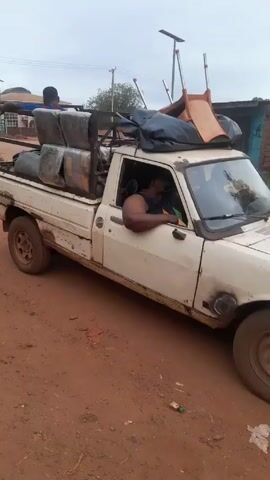 A white, heavily loaded pickup truck is seen moving along a red dirt road. The truck bed is packed with what appears to be bales of material wrapped in plastic, as well as chairs and other items secured with a tarp. A person is sitting atop the load in the truck bed, appearing to be holding on. The driver is visible through the truck's open window. The setting appears to be a rural or semi-rural area with trees and buildings in the background.