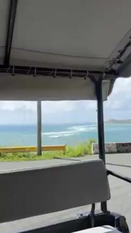 The video, shot from inside a covered golf cart, shows a road winding along a coastline on a bright day with a partly cloudy sky. The road has a yellow guardrail on the ocean side, separating it from green vegetation and the turquoise ocean. There are power lines running alongside the road.  The video captures the movement of the golf cart as it drives along the road.