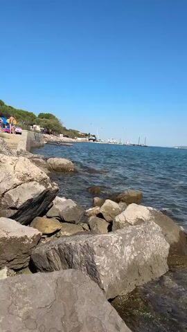 The video begins by showing a rocky shoreline with the clear blue water of the sea lapping at the rocks. The camera pans to the right, revealing a large cargo ship in the distance, as well as a city or town visible along the coastline. The water is clear enough to see the pebbles and rocks beneath the surface. The camera continues to pan, showing more of the coastline, including hills in the background. A small inflatable palm tree floats in the water. The sky is clear and blue throughout the video. There are no people clearly visible in the foreground, but a few people can be seen swimming in the water in the distance. The video appears to have been filmed on a sunny day.
