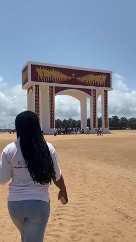 The video shows a woman walking towards a large monument resembling an archway, set in a sandy, open space under a partly cloudy sky. The archway is white with a maroon trim and features golden figures in relief across the top. A group of people is gathered near the monument. The woman is wearing a white t-shirt and jeans, and her hair is styled in braids. She is holding a cell phone in her hand. The ground appears to be sandy, with some vegetation visible in the background. The overall scene suggests a tourist or public gathering at a cultural or historical site.