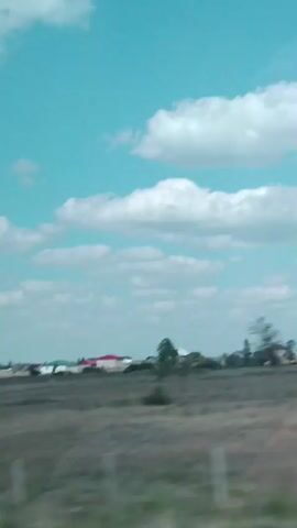 The video shows a panoramic view of a rural landscape under a bright blue sky with scattered white clouds. In the foreground, there is a dry, grassy field. In the middle ground, a few buildings, possibly houses and construction sites, are visible, interspersed with trees and vegetation. The buildings have various colored roofs, including red and green. The camera appears to be moving, as indicated by the slight blur and the changing perspective of the landscape. The overall impression is that of a quiet, undeveloped area.