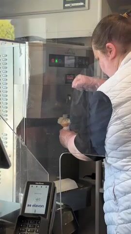 This media file captures a person making ice cream in what appears to be a cafe or food establishment. The person, wearing a white puffer vest and clear gloves, is operating an ice cream machine, holding a cone beneath its dispenser. The machine is labeled "CARPIGIANI" and has illuminated buttons. In the foreground, a Verifone payment terminal displays "elavon" and a welcoming message. The scene is indoors, with a bright, likely daytime setting as suggested by the lighting and a glimpse of greenery through a window. The overall atmosphere seems to be one of a typical service interaction. There are no strong emotional cues visible, but the activity suggests a moment of food preparation and service. The location is identified as Westport, Ireland.