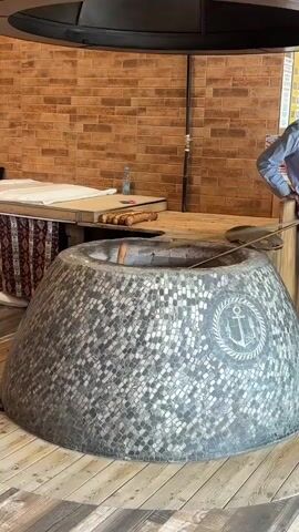 The video shows a tandoor oven, cylindrical and decorated with gray and white mosaic tiles. An anchor design is emblazoned on the side of the oven. A wooden table is visible behind the oven, holding a stack of what appear to be loaves of bread, and a water bottle. A person can be seen in the background next to the table, but their actions are unclear. The environment suggests a kitchen or restaurant, indicated by the brick wall in the background. A metal tool lies in the oven.