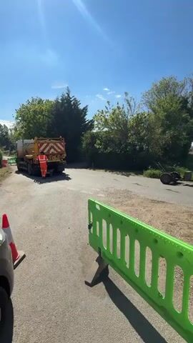 The video shows a road work site on a sunny day. Green and red-and-white safety barriers surround an area with broken asphalt, purple cable spools, and other equipment. Workers are present, with one seen unloading materials from a truck. Another truck with a lift is parked nearby. The site is located on a road next to a field.