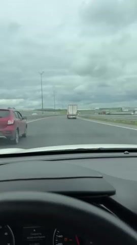 The view from the interior of a vehicle driving on a highway, looking towards the road ahead, is captured in this video. Another vehicle, a red SUV, is in the adjacent lane on the left, and a white truck is farther ahead in the same lane as the camera vehicle. The sky is overcast with grey clouds. Roadside lighting is visible on the left, and the highway appears to be elevated over other roads. The dashboard and steering wheel of the camera vehicle are visible in the foreground. The video captures the movement of the car along the highway under a cloudy sky.