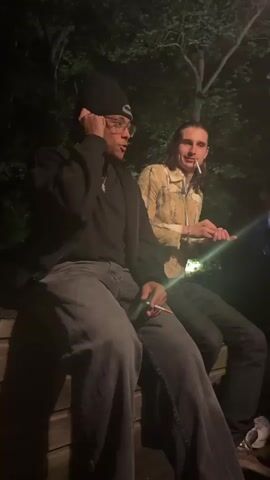 Two young men are seated outdoors at night. The person on the left wears a black beanie, glasses, a black top, and grey jeans. He puts his hand up to his head and appears to be talking on the phone. He is holding a cigarette in his hand. The person on the right is wearing a light patterned jacket and dark jeans. He is holding a cigarette in his mouth. A third person enters the frame and lights the first person's cigarette. There are trees in the background.