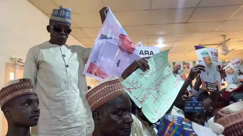 A group of men stand in a room, holding up signs. The signs feature the same man's image, and one sign is handwritten saying "WE NEED INTERVENTION OF APC NATIONAL LEADER". Most of the men are wearing traditional hats and clothing. There are several cameras in the room, suggesting a press conference or political rally. The room has white walls and a drop ceiling.
