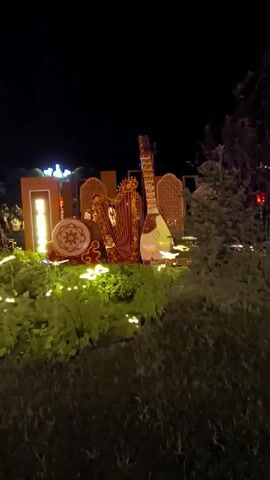 The video pans across a nighttime outdoor scene decorated with lights. In the foreground, a display features illuminated musical instruments, including a harp and a lute-like instrument, along with a drum. The instruments are intricately decorated and appear to be part of a larger decorative arrangement. The area around the display is filled with small, scattered lights, creating a festive atmosphere. In the background, trees and foliage are visible, also illuminated by strings of lights. Further in the distance, a tall structure with multiple lights and red lanterns can be seen, as well as the lights of buildings. The overall impression is that of a nighttime outdoor event or display, possibly a festival or holiday celebration. There are no people visible in the video.