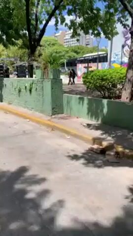 The video shows a street scene. The camera pans from a green-painted wall to a taxi driving on the street. The camera then continues moving down the street, past buildings with graffiti on the walls. A truck can be seen further down the road. The day is sunny with a blue sky and some clouds. Trees are visible along the street.